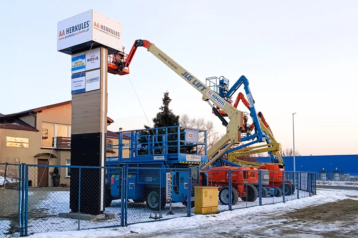 Kilka podnośników koszowych obok słupa reklamowego na ogrodzonym, ośnieżonym placu.