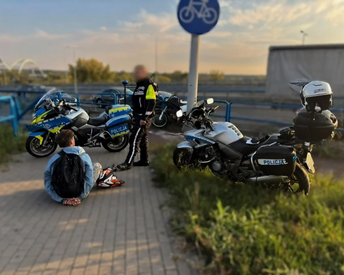 Funkcjonariusz policji motocyklowej stoi przy policyjnych motocyklach obok mężczyzny siedzącego na chodniku na ścieżce rowerowej.