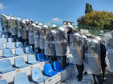 Funkcjonariusze polskiej policji w hełmach i z tarczami ustawieni w szeregu na trybunach stadionu.