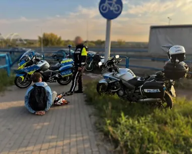 Funkcjonariusz policji motocyklowej stoi przy policyjnych motocyklach obok mężczyzny siedzącego na chodniku na ścieżce rowerowej.