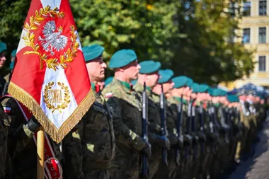 Żołnierze polscy w zielonych beretach i mundurach kamuflażowych stoją w szeregu z bronią podczas uroczystej formacji na otwartej przestrzeni miejskiej, a na pie