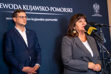 Kobieta w szarym garniturze przemawia do mikrofonów podczas konferencji prasowej w siedzibie urzędu wojewódzkiego kujawsko-pomorskiego, obok stoi mężczyzna w gr