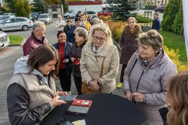 Grupa osób stoi przy okrągłym stole na brukowanej nawierzchni na tle parkingu i zieleni, jedna kobieta obsługuje urządzenie mobilne.