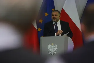 Mężczyzna w garniturze przemawia z pulpitu z orłem, za nim flagi Unii Europejskiej i Polski.