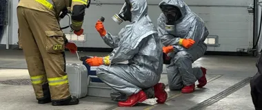 Strażak i dwie osoby w srebrnych kombinezonach ochronnych z maskami gazowymi badają próbkę przy użyciu sprzętu pomiarowego w pomieszczeniu garażowym.