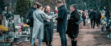 Grupa osób stoi na cmentarzu przy grobach ozdobionych kwiatami i zniczami, rozmawiając i pijąc z kubków.